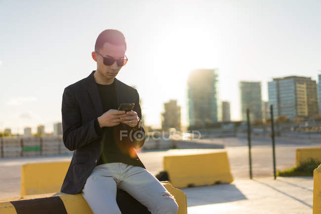 Junger Asiate mit Sonnenbrille benutzt Smartphone auf Parkplatz — Stockfoto