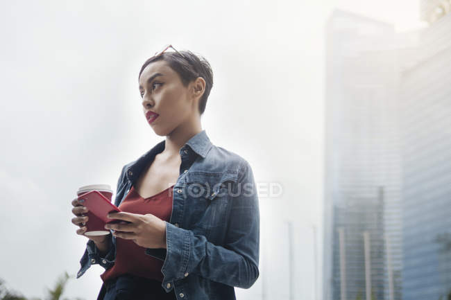 Jovem senhora malaia cingapuriana em um ambiente urbano com seu smartphone e uma xícara de café nas ruas . — Fotografia de Stock