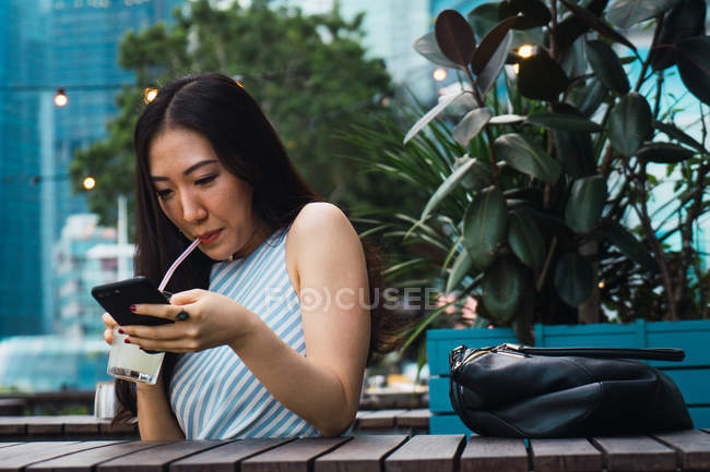 Jovem mulher asiática tendo bebida e usando smartphone — Fotografia de Stock