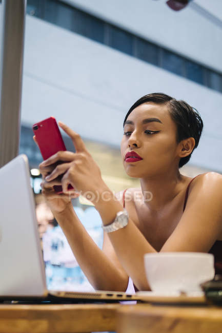 Halbporträt einer jungen malaiischen Singaporeanerin, die während der Arbeit ihr Handy in einem Café benutzt. — Stockfoto
