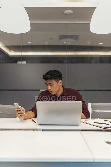 Молодой азиатский бизнесмен, работающий с ноутбуком и смартфоном в современном офисе — стоковое фото