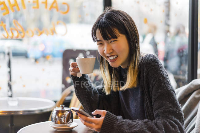 Молода приваблива випадкова азіатка зі смартфоном і кавою в кафе — стокове фото