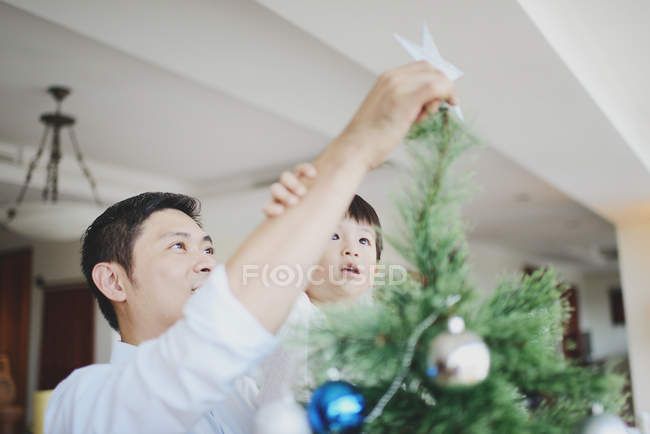 Азиатская семья празднует Рождество, отец с сыном украшают елку — стоковое фото