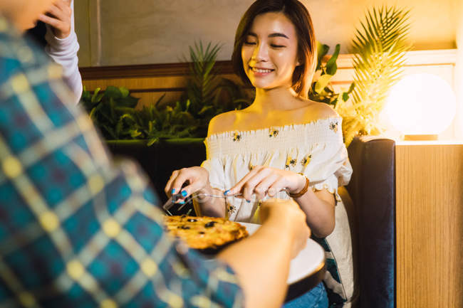 Giovane donna asiatica affettare la pizza in confortevole bar — Foto stock