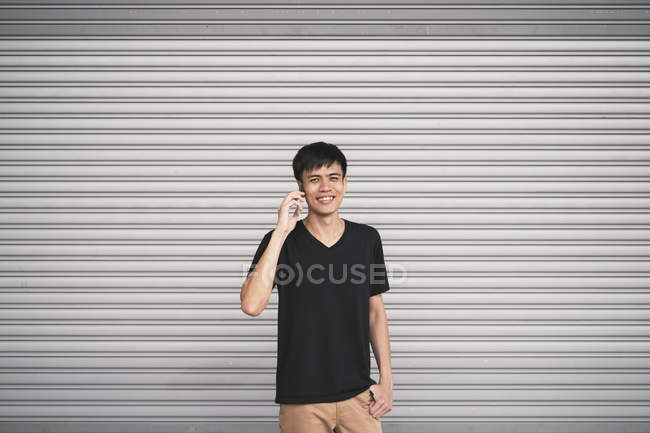 Jeune homme asiatique avec téléphone mobile posant contre le mur gris — Photo de stock