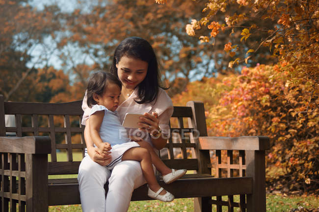 Bonito asiático mãe e filha usando smartphone no banco — Fotografia de Stock