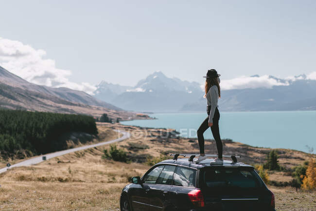 Mujer joven sentada encima del coche en Milford Sound, Nueva Zelanda - foto de stock