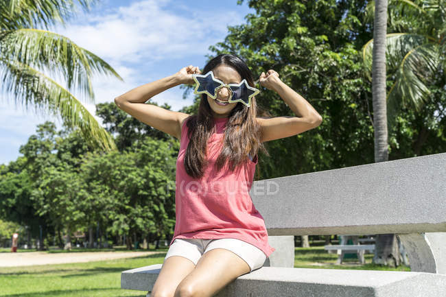 Счастливая азиатская взрослая женщина сидит на скамейке и позирует в смешных солнцезащитных очках — стоковое фото