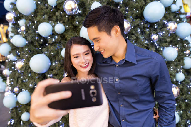 Jeune attrayant asiatique couple ensemble shopping dans le centre commercial à Noël et de prendre selfie contre sapin — Photo de stock