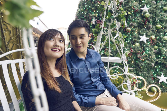 RELEASES Happy young asian couple sitting on swing near fir tree — Stock Photo