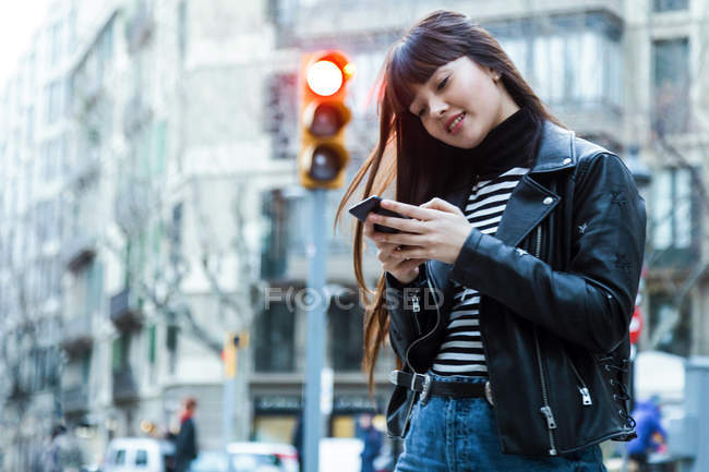 Jeune femme aux cheveux longs marchant et naviguant sur son smartphone — Photo de stock