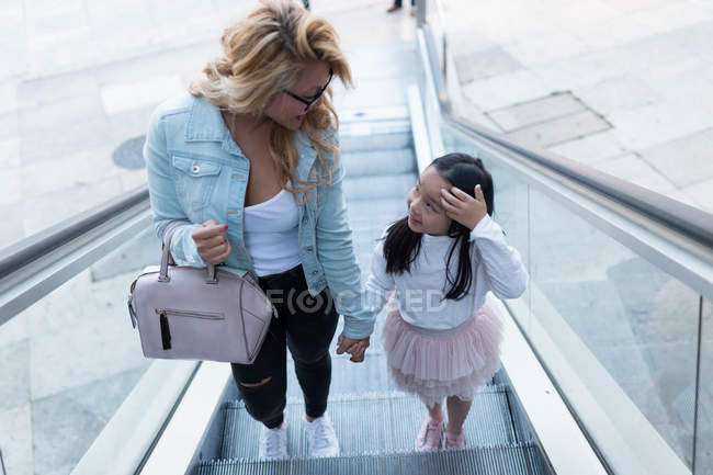 Glückliche junge Mutter mit ihrer Tochter im Gespräch auf der Rolltreppe in der Stadt. — Stockfoto