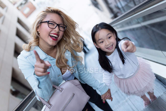 Feliz joven madre con su hija en la escalera mecánica señalando - foto de stock