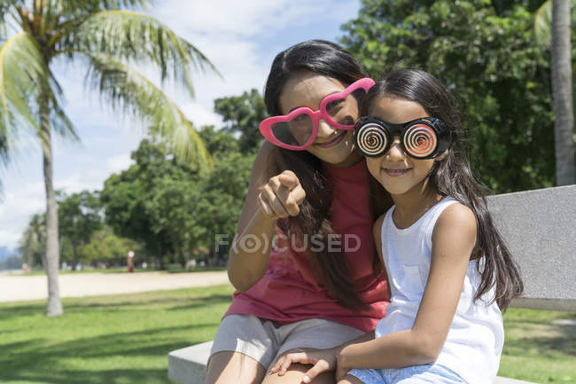 Jovem asiático mãe com bonito filha sentado no banco no engraçado óculos — Fotografia de Stock