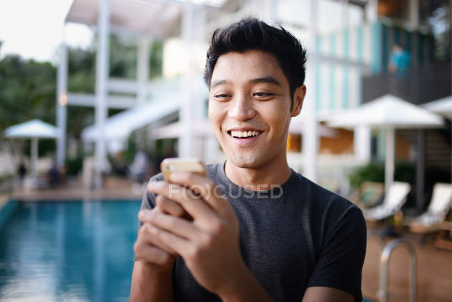 Giovane attraente asiatico utilizzando smartphone contro piscina — Foto stock