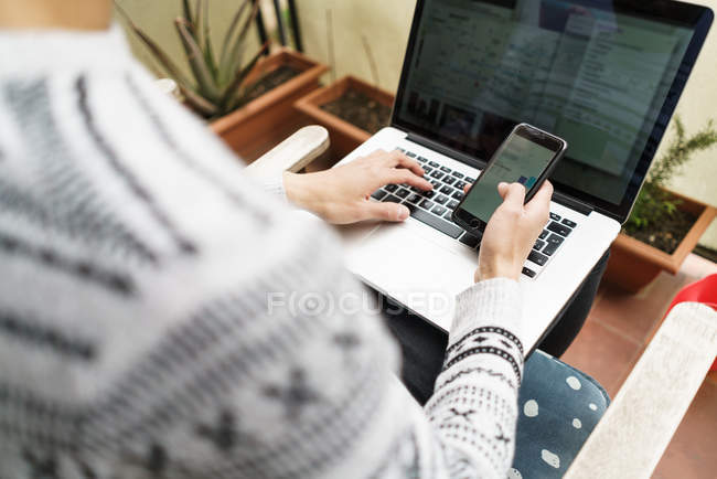 Imagen recortada de hombre adulto joven usando el ordenador portátil y el teléfono inteligente en casa - foto de stock