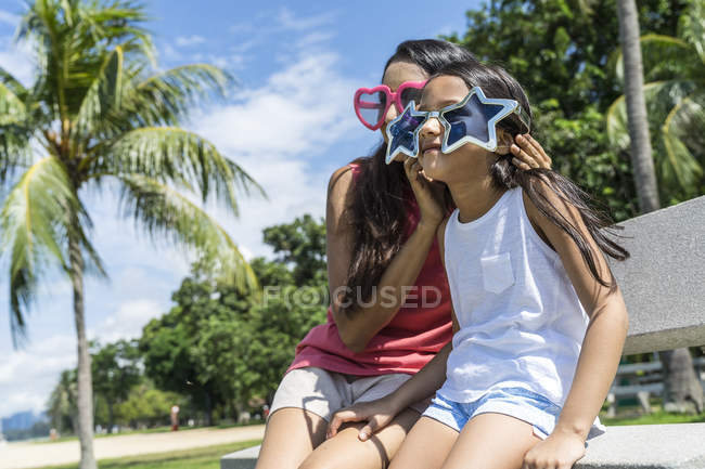 Молода азіатська мати з милою дочкою сидить на лавці в кумедних окулярах — стокове фото