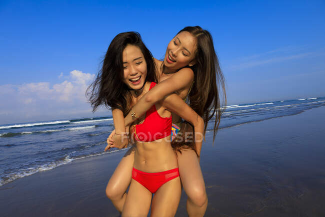 Dois jovens asiáticos amigos do sexo feminino estão brincando na praia. Um está tomando teh outro em suas costas e carregando-a rindo. — Fotografia de Stock