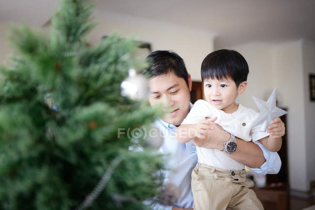 Счастливая азиатская семья празднует Рождество вместе дома, отец и сын украшая елку — стоковое фото