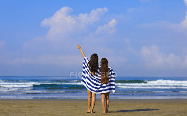 Dos amigas con el pelo largo están de pie en una playa envuelta en una toalla de rayas azules y blancas disfrutando de la vista al mar . - foto de stock