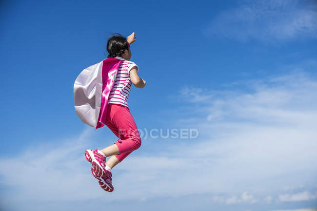 Cute little superhero girl in mask and coat jumping against sky — Stock Photo