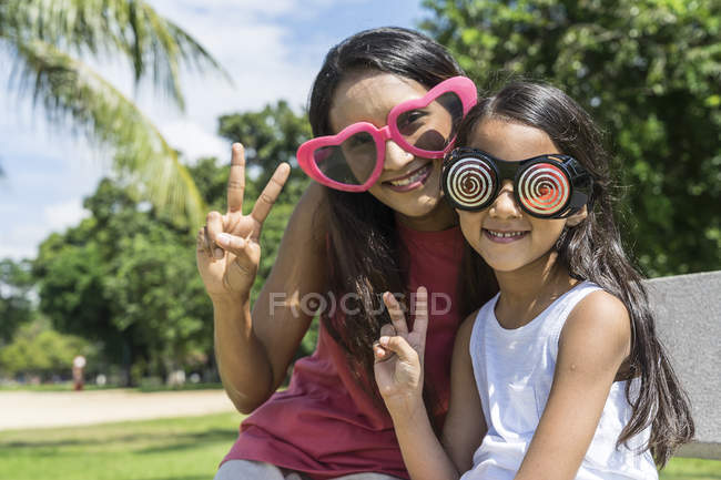 Молодая азиатская мать с симпатичной дочерью сидит на скамейке в смешных очках — стоковое фото
