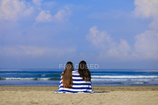 Dos amigas con el pelo largo están sentadas en una playa adornada con pegamento y una toalla blanca a rayas disfrutando de la vista al mar. - foto de stock
