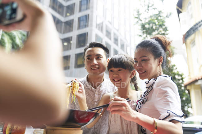 Happy asian family eating noodles together in street cafe and taking photo — Stock Photo