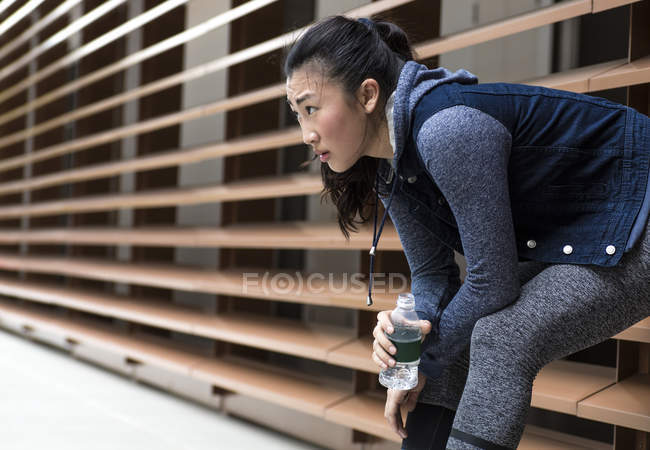 Una joven asiática está descansando en una pared después de su entrenamiento corriendo por su vecindario. Ella sostiene una botella de agua . - foto de stock