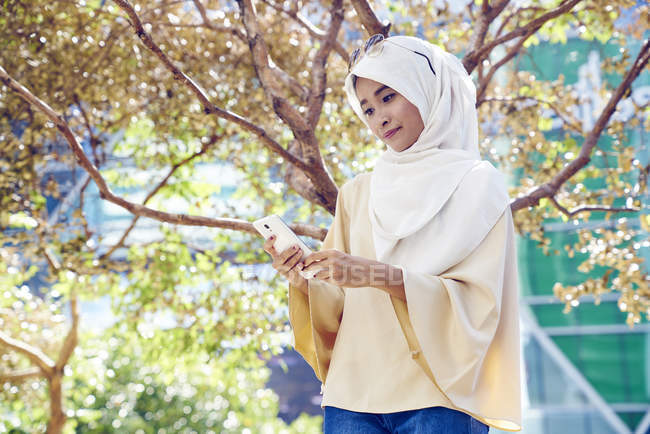 Schöne malaiische Frau im Hidschab im Park mit ihrem Handy — Stockfoto