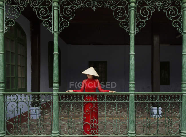 Mujer vietnamita con sombrero cónico de pie en un balcón en Hoi An en su Ao Dai - foto de stock