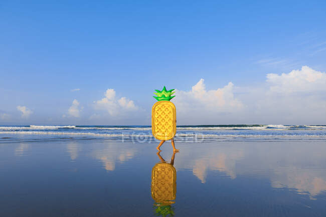 Un ananas galleggiante è a piedi lungo la spiaggia di Bali. Una persona lo sta portando, mostrando solo le gambe. — Foto stock