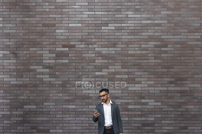 Handsome indian business man using smartphone against brick wall — Stock Photo