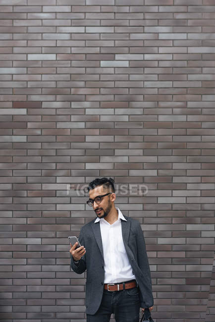 Homem de negócios indiano bonito usando smartphone contra parede de tijolo — Fotografia de Stock