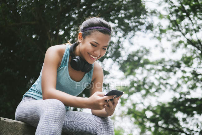 Молодая азиатская спортсменка с помощью смартфона в парке — стоковое фото
