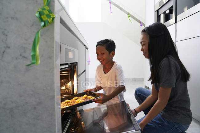 Jóvenes hermanos asiáticos celebrando Hari Raya juntos en casa y cocinando platos tradicionales - foto de stock