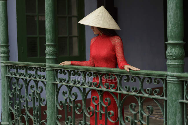 Mujer vietnamita con sombrero cónico de pie en un balcón en Hoi An en su Ao Dai - foto de stock
