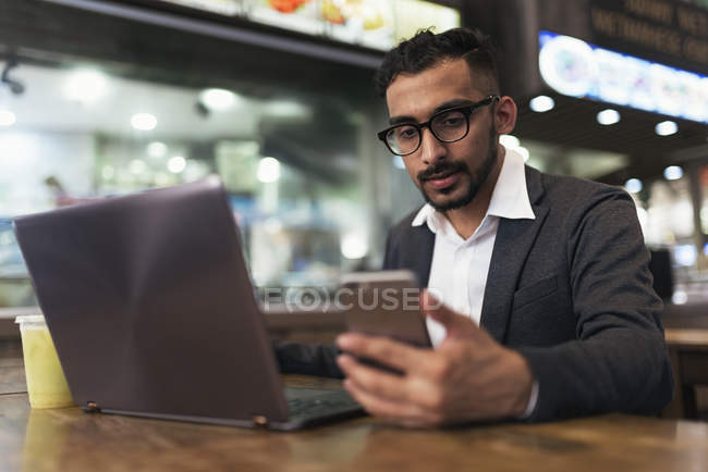 Handsome indian business man using smartphone and laptop in cafe — Stock Photo