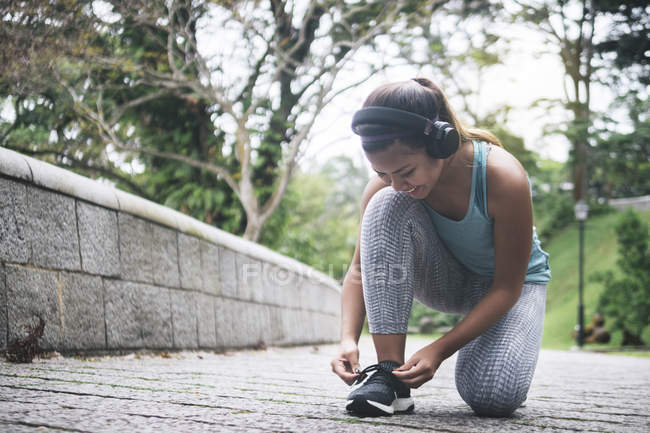 Молода азіатська спортивна жінка в навушниках зв'язуючи шнурки в парку — стокове фото