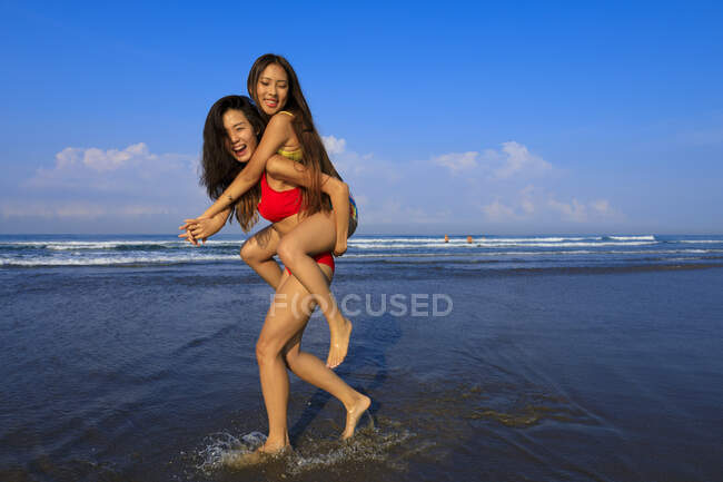 Deux jeunes amies asiatiques s'amusent à la plage. L'une prend l'autre sur son dos et la porte en riant — Photo de stock