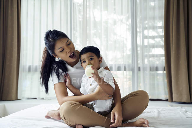 Asiático madre relojes como su hijo se alimenta de leche botella - foto de stock