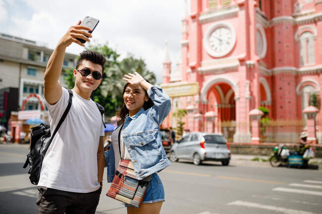 Joven pareja asiática tomando selfie contra catedral, Ho Chi Minh City, Vietnam - foto de stock