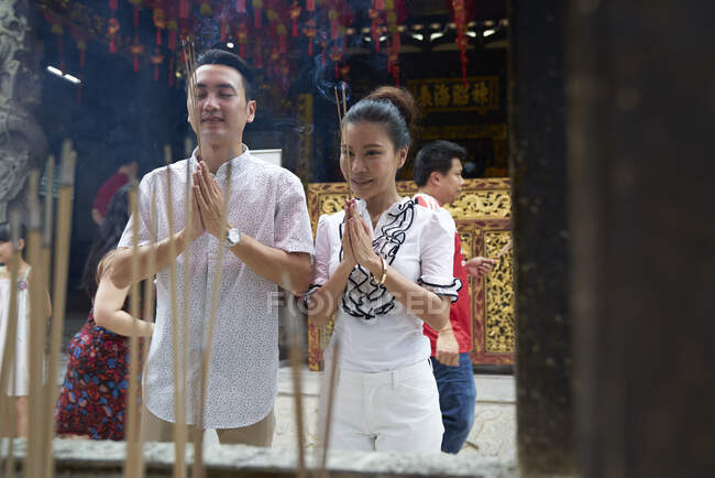 Joven asiático hombre y mujer orando en templo con joss palos - foto de stock