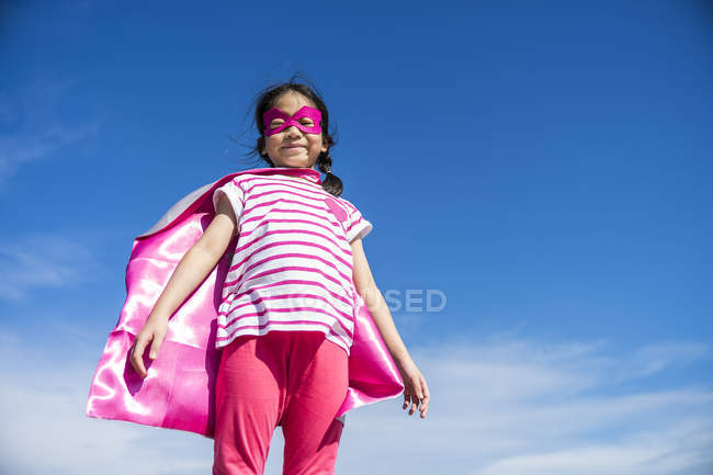 Linda niña superhéroe en máscara y abrigo posando contra el cielo - foto de stock