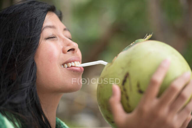 A young pretty woman is sipping on a freshly opened coconut with a smile on her face — Stock Photo