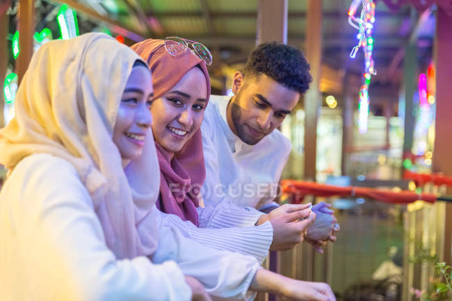 Three muslim friends looking down at mobile phone on a bridge at night time, while one girl looks into the camera — Stock Photo