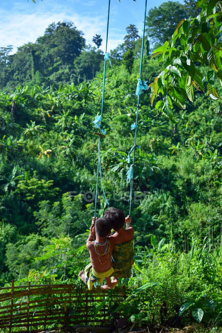 Deux enfants chakma se balançant dans une balançoire faite par eux-mêmes au bord de la montagne, chantant une de leurs chansons religieuses — Photo de stock