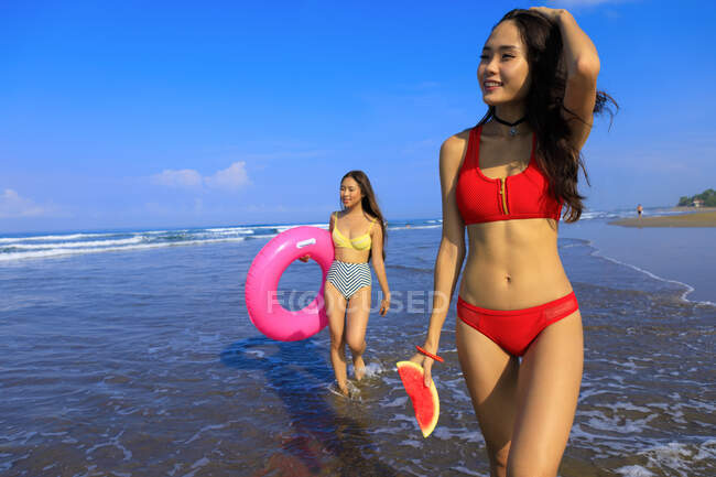 Zwei hübsche Mädchen mit Schwimmweste und Wassermelone laufen am Ufer entlang. Sie haben eine gute Zeit. — Stockfoto