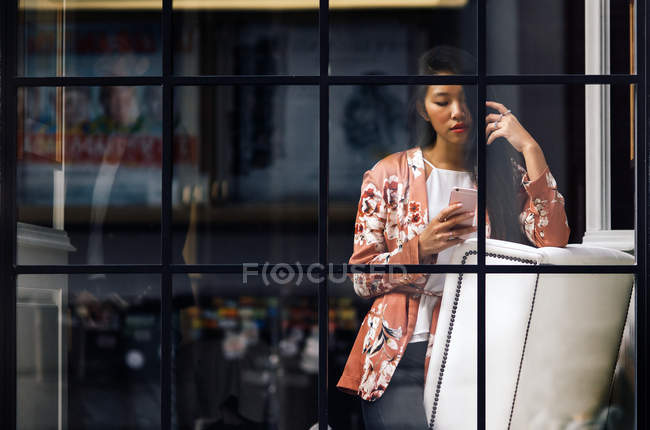 Piuttosto lungo capelli donna cinese utilizzando smartphone attraverso la finestra — Foto stock
