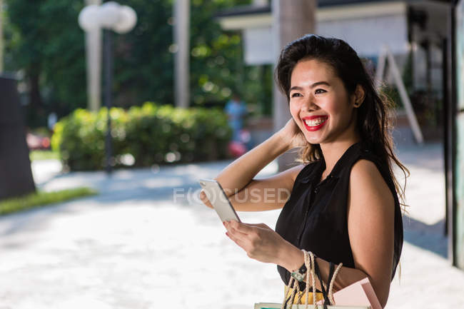 Sonriendo mujer asiática con bolsas de compras usando teléfono inteligente - foto de stock